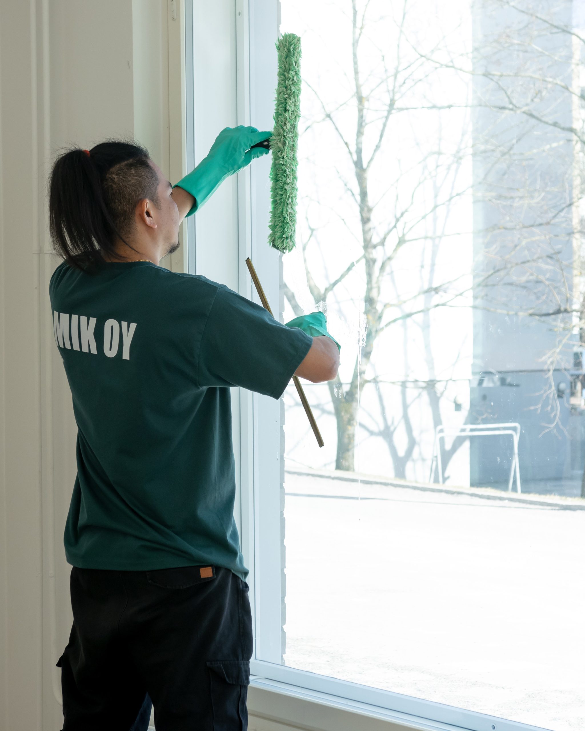 Mik Oy branded crew member in green uniform cleaning a commercial office window interior with professional tools during winter in Helsinki, Finland