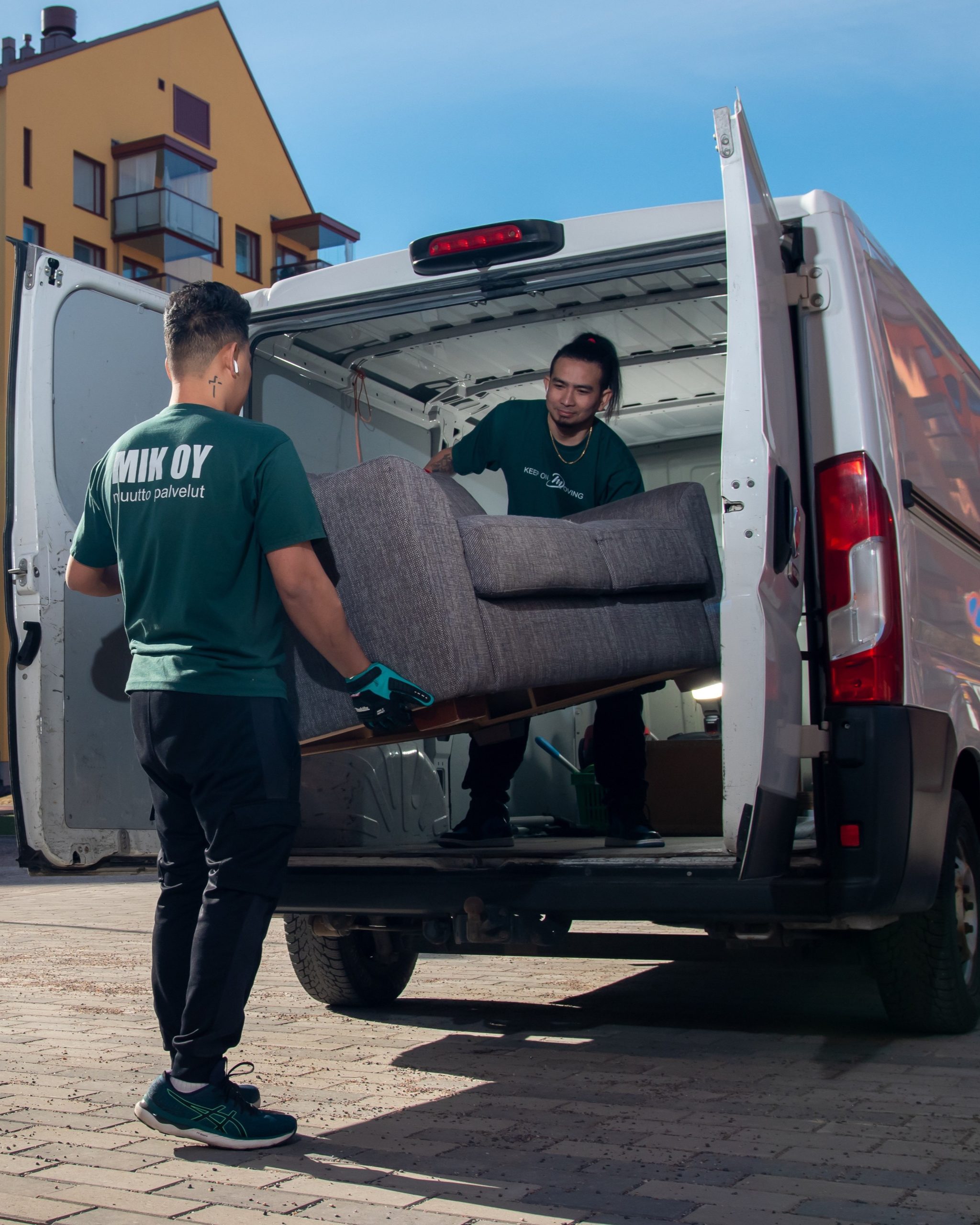 Kaksi Mik Oy:n työntekijää vihreissä MIK OY -työasuissa lastaa harmaata sohvaa pakettiautoon yrityskuljetuksessa Helsingissä