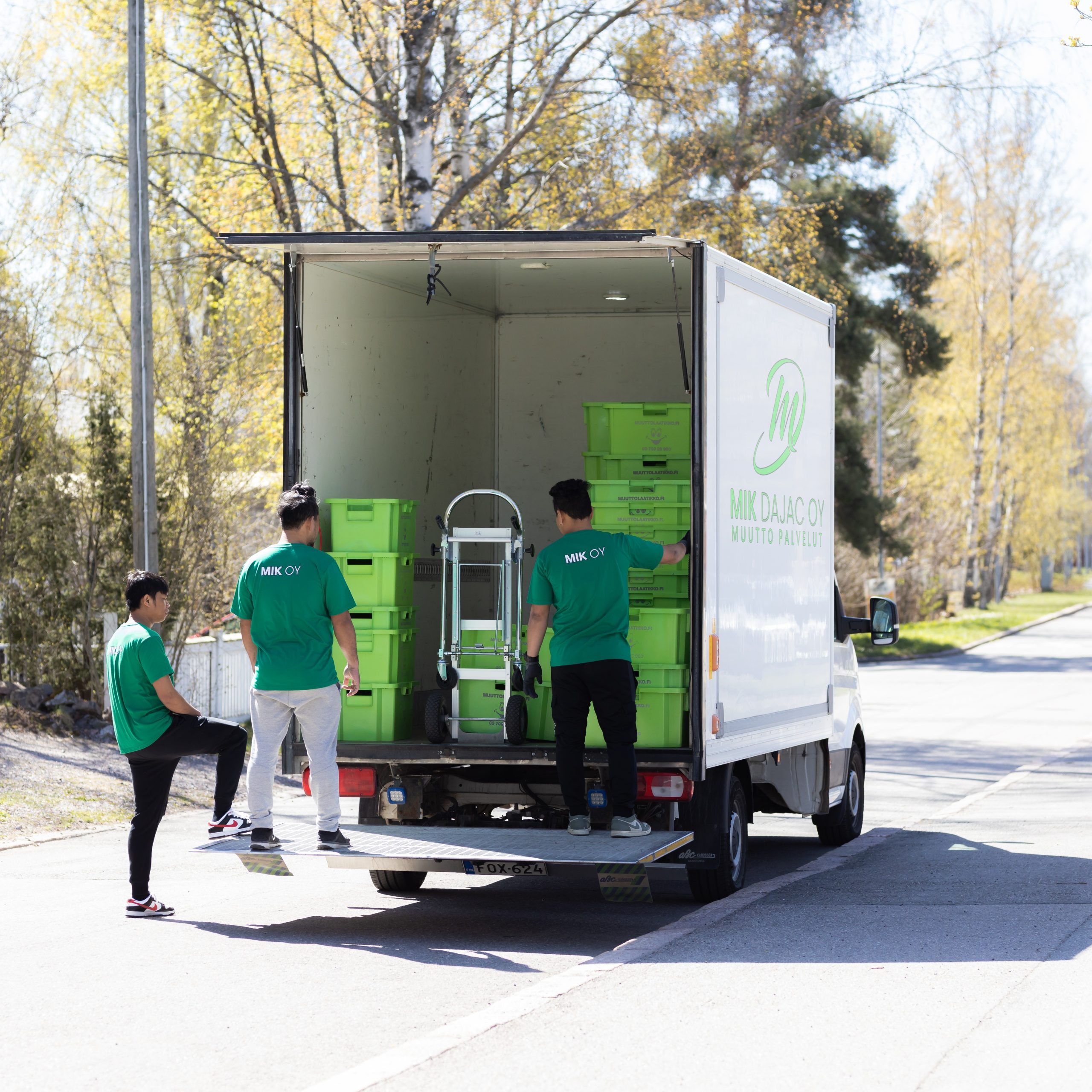 Three Mik Oy crew members in green uniforms loading green moving crates into a branded Mik Dajac Oy Muutto Palvelut truck during a residential move in Lahti, Finland