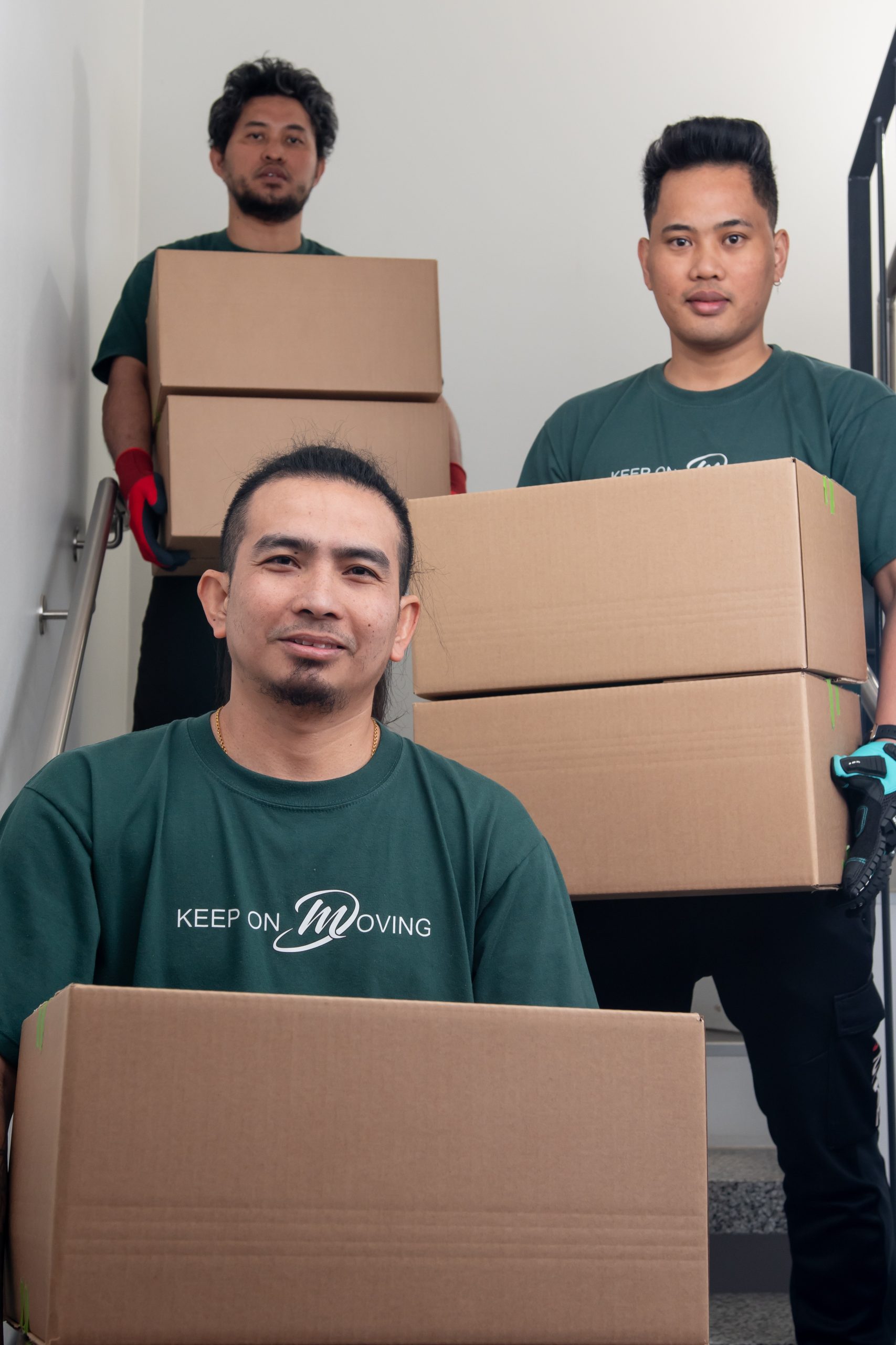 Three Mik Oy crew members in green uniforms carrying moving boxes down a residential staircase during a home move in Lahti, Finland