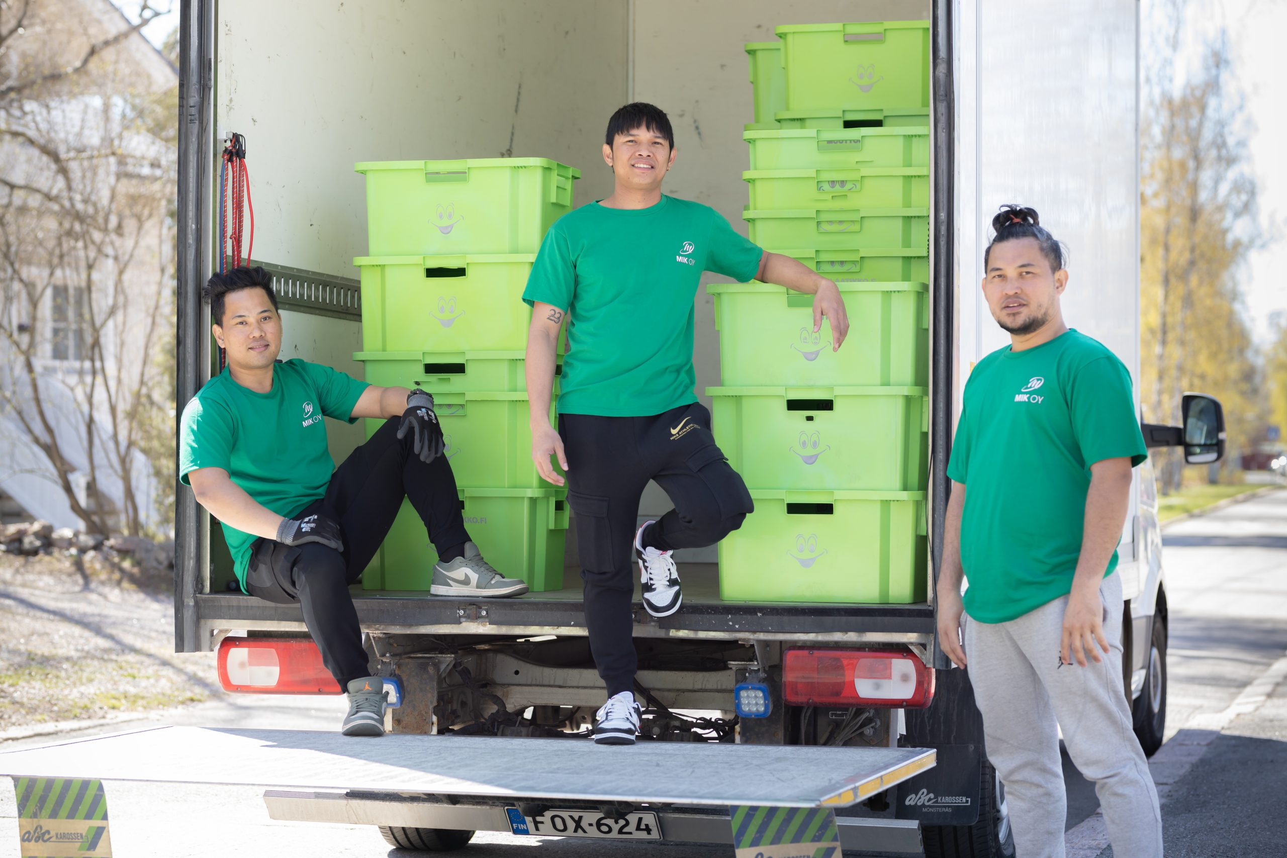 Three Mik Oy crew members in green uniforms posing confidently at the back of a fully loaded moving truck with green muuttolaatikko crates in Lahti, Finland